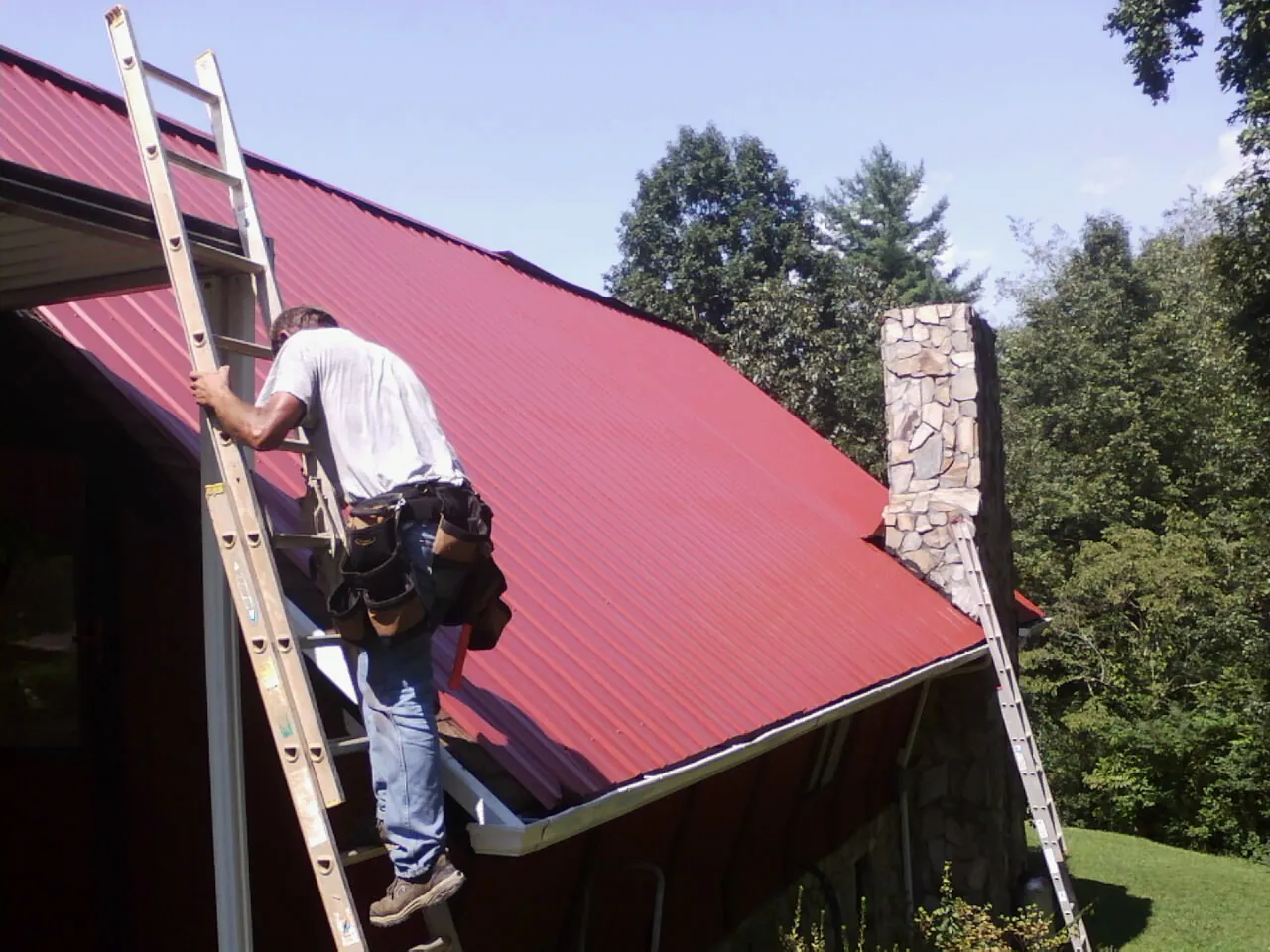 Roofer on a ladder beside a red metal roof and stone chimney — Soffit & Fascia Repair by Roofing Brothers in Camino